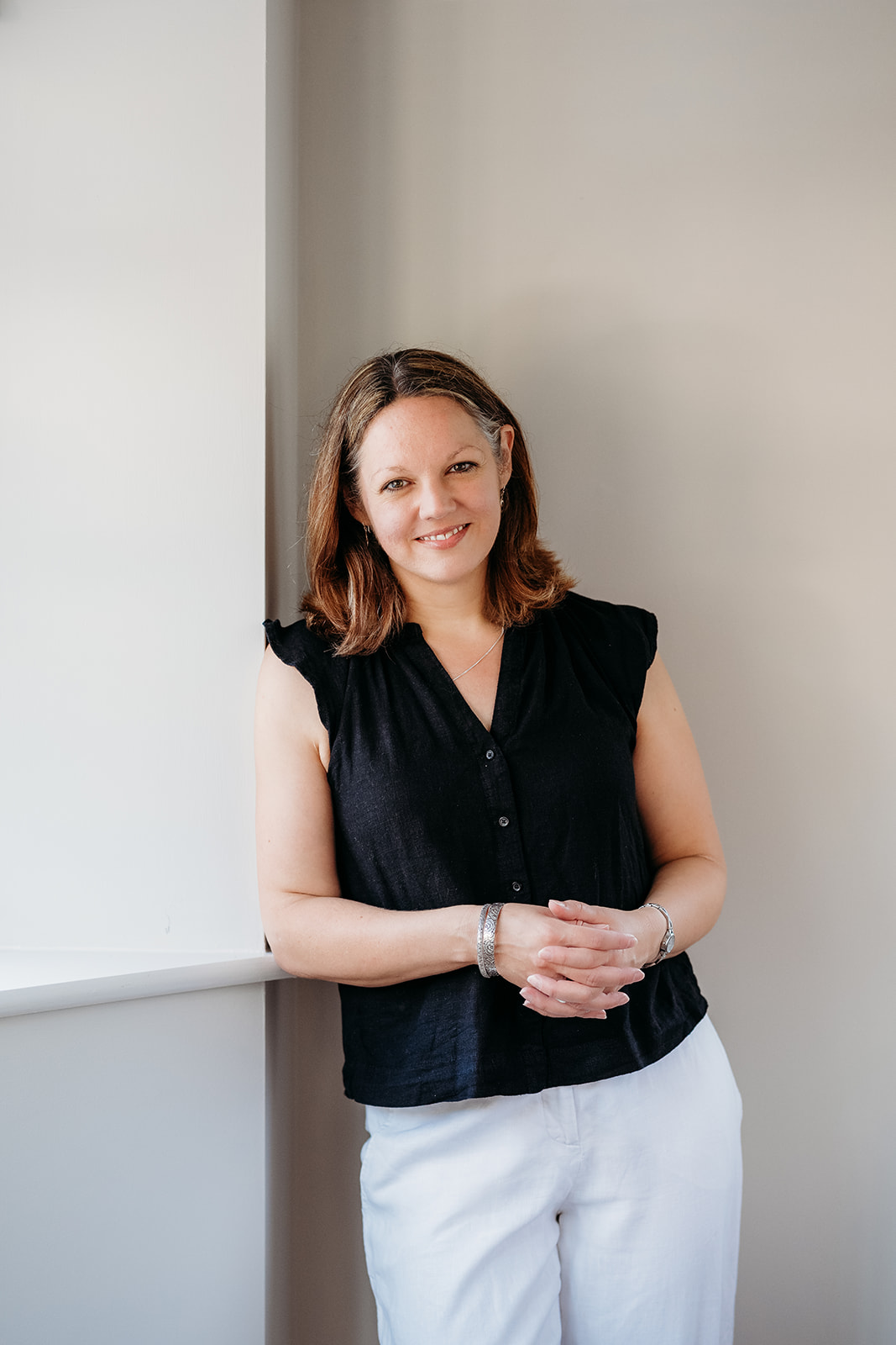 A woman with shoulder-length brown hair smiles while leaning against a wall, wearing a black sleeveless top and white pants.