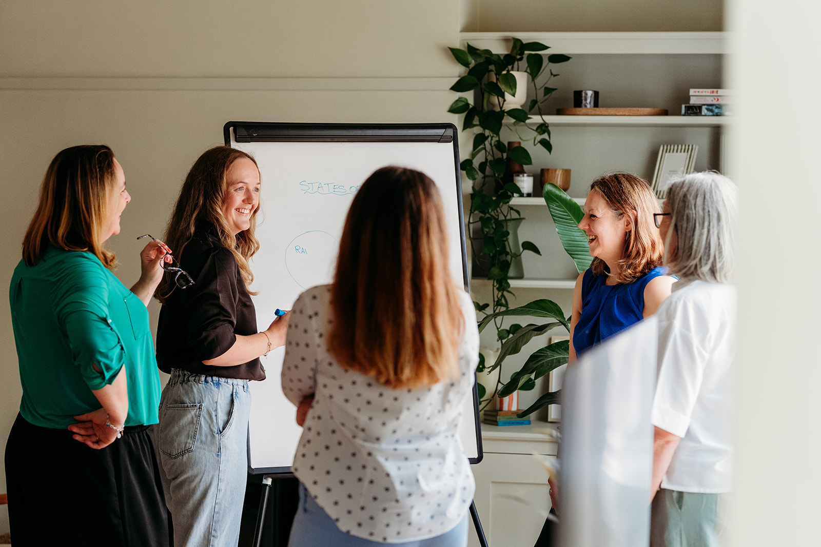 Four women gathered around a whiteboard in a bright room, discussing work and smiling.